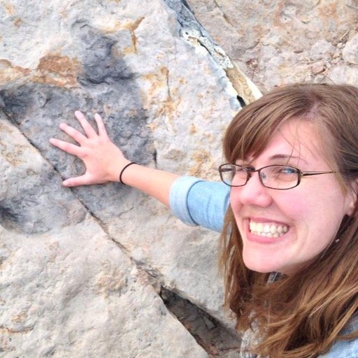 White woman with glasses smiles at camera. hand outstretched onto a dinosaur footprint 
